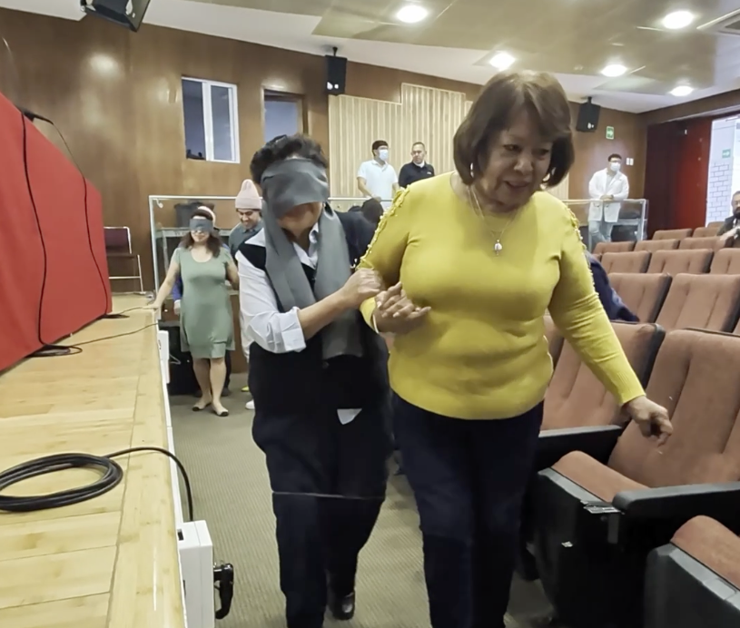 Dos personas realizan una actividad, una de ellas tiene los ojos vendados y las piernas están atadas con un cordón, la otra persona le apoya para sostenerla con su brazo y permitir que camine con su apoyo