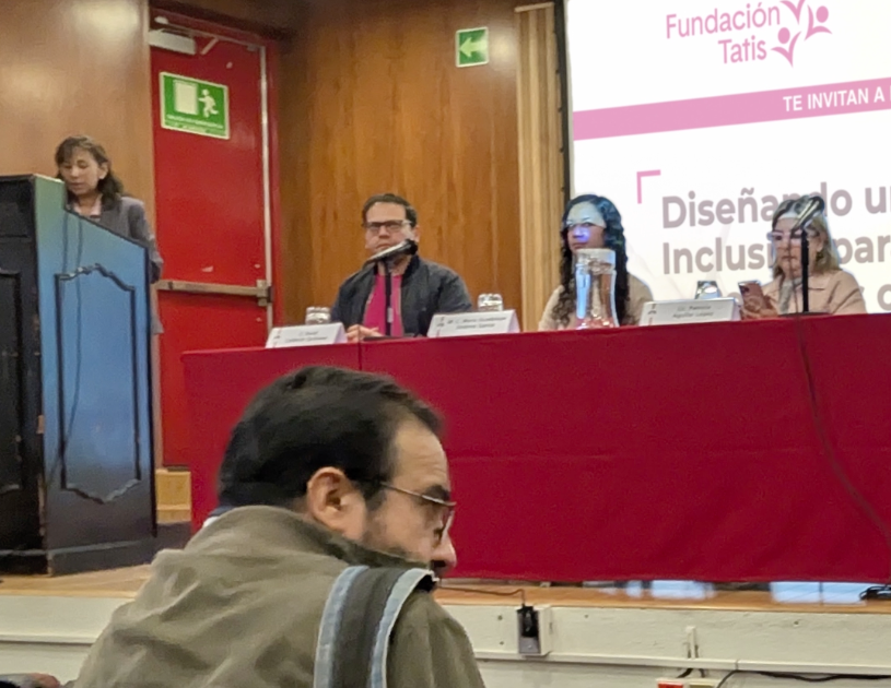 Al frente de un auditorio esta un podium a la izquierda, una persona ponente se dirige al público, a la derecha en una mesa sentados están las personas ponentes, al fondo una proyección sobre una pantalla en la que se observa el logotipo de Fundación Tatis