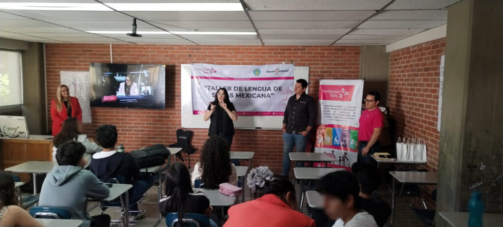 Dos personas instructoras se dirigen a personas asistentes, al fondo una lona que dice Taller de Lengua de Señas Mexicana y los logos de Fundación Tatis y Voluntariado Columbia