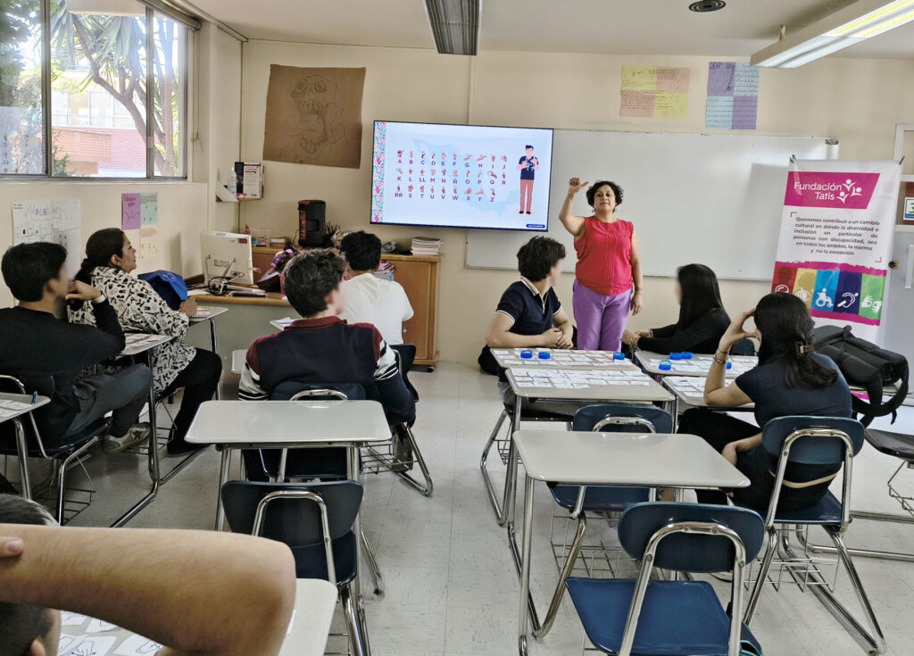 Persona instructora al frente de un salón explica la interpretación de una Señas a personas asistentes, al fondo en una pantalla esta proyectado el abecedario en Lengua de Señas Mexicana