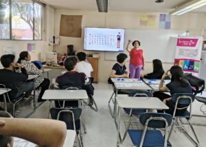 Persona instructora al frente de un salón explica la interpretación de una Señas a personas asistentes, al fondo en una pantalla esta proyectado el abecedario en Lengua de Señas Mexicana